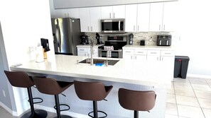 Eine moderne Küche mit einem Edelstahlkühlschrank, einer Mikrowelle und einer Kaffeemaschine. Unter einer weißen Kücheninsel stehen drei braune Barhocker.