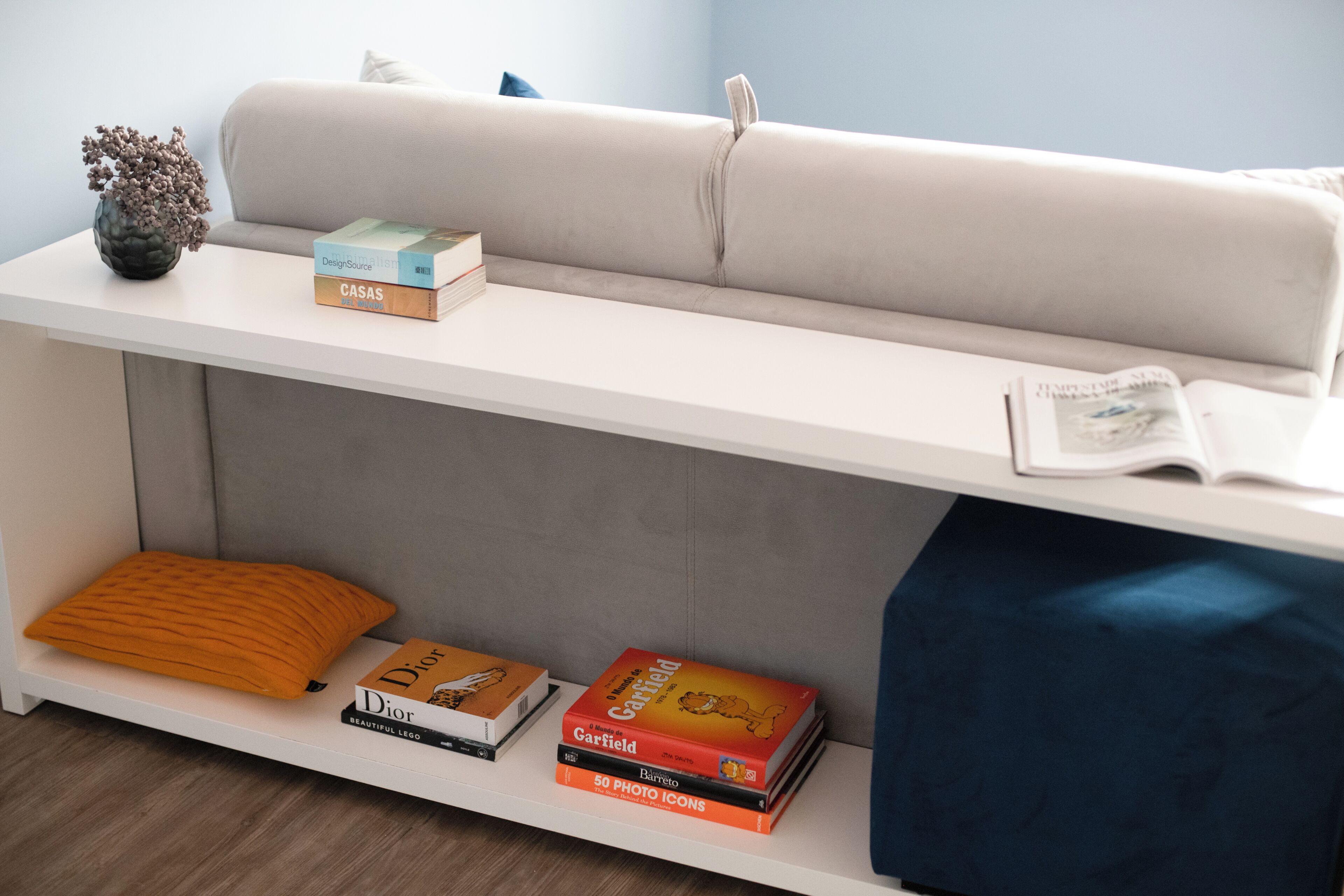 Um sofá branco com uma almofada, uma mesinha com livros, um vaso com flores e uma almofada azul no chão.