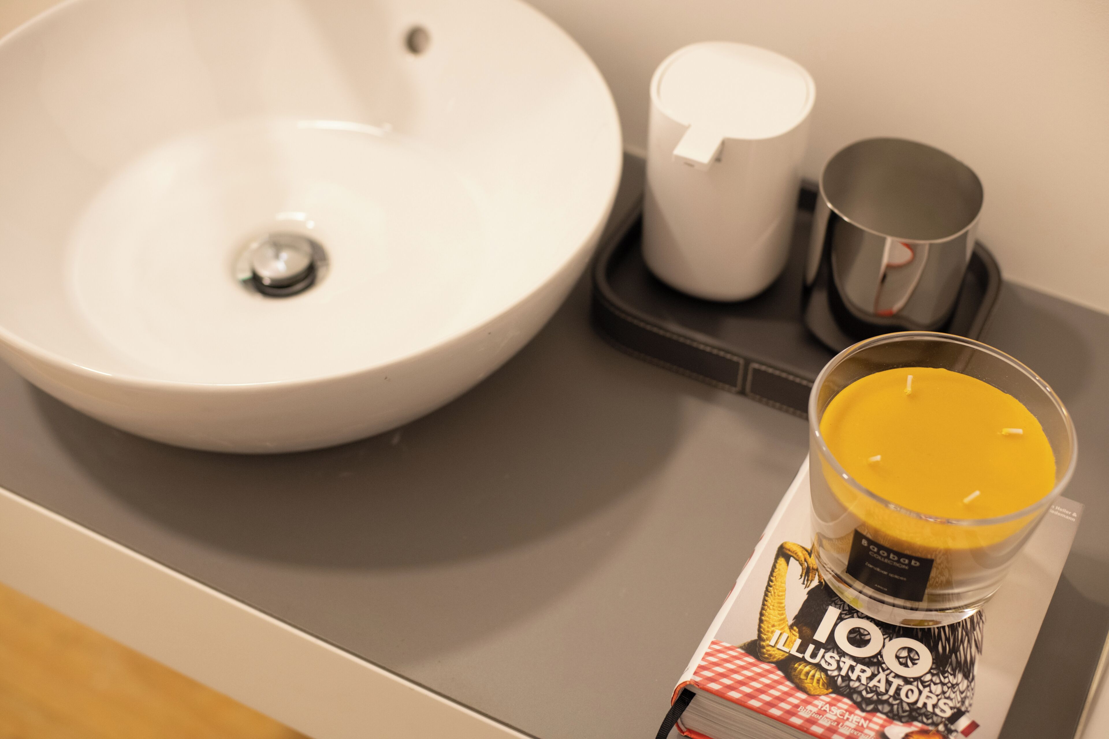 A bathroom sink, a white cup, a metallic mug, and a candle in a glass holder.