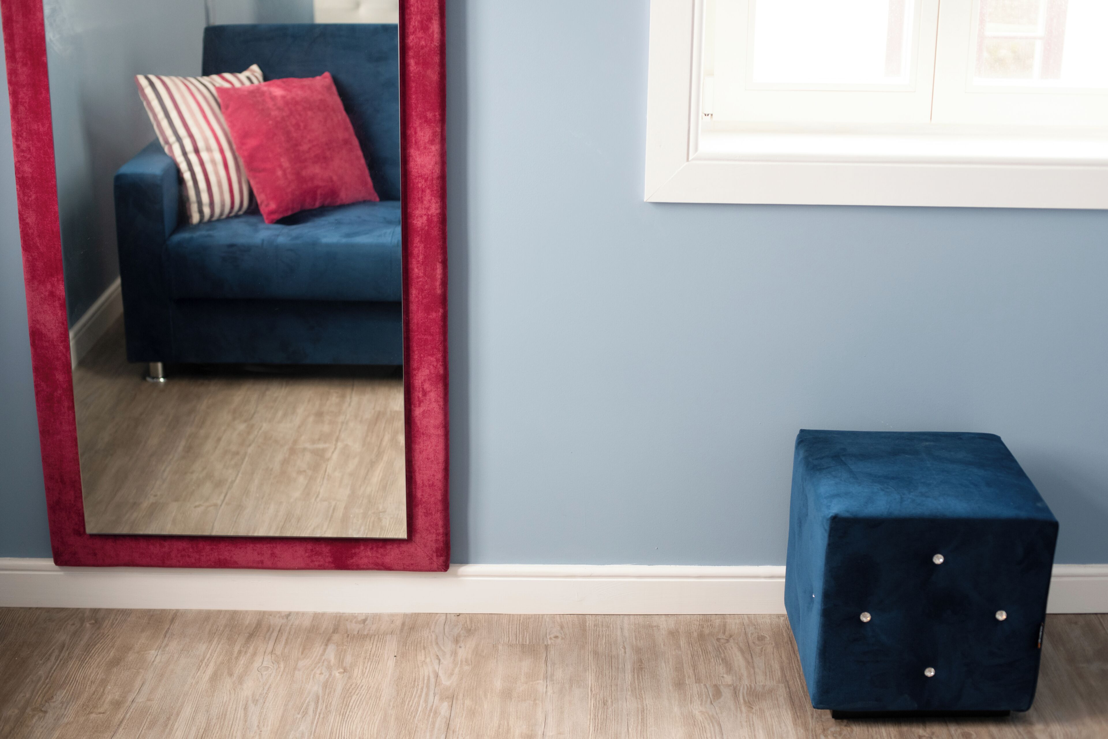 Ein Zimmer mit einem blauen Sofa, einem spiegel mit rotem Rahmen und einem dunkelblauen Pouf.