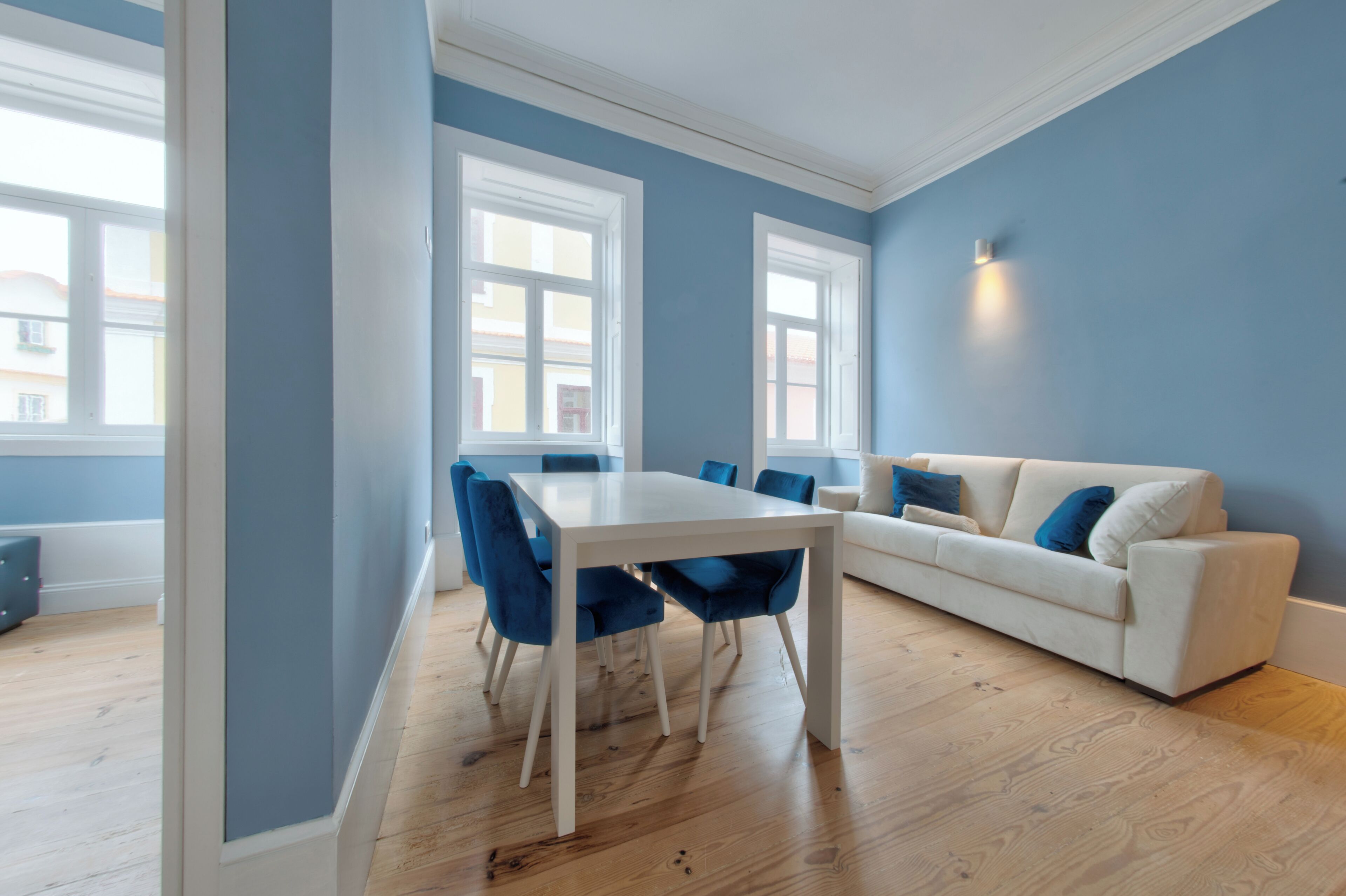 A modern living room with a white sofa, blue chairs, a white dining table, and large windows.