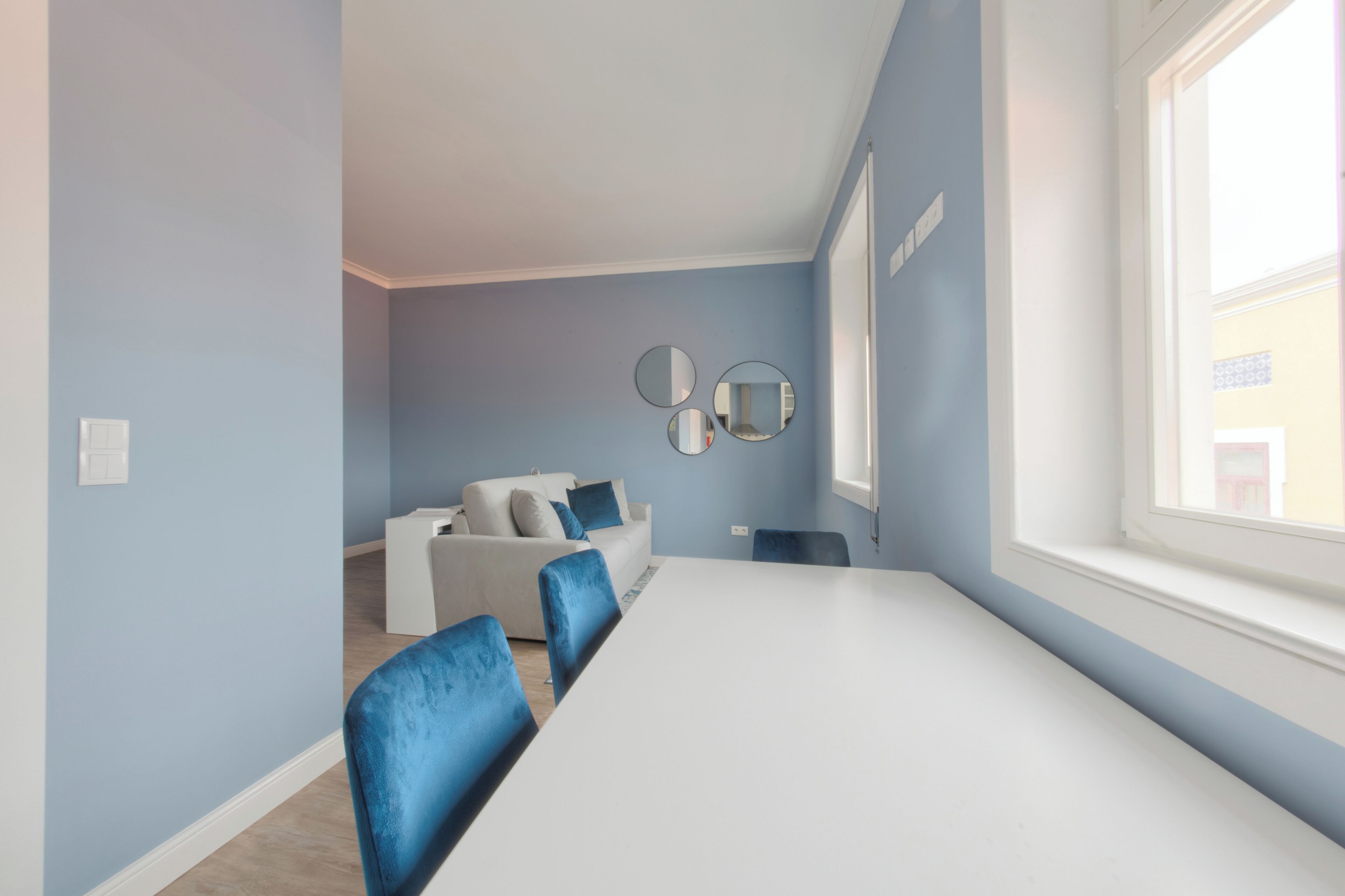 Una habitación con una mesa blanca, sillas azules, un sofá blanco y una ventana.