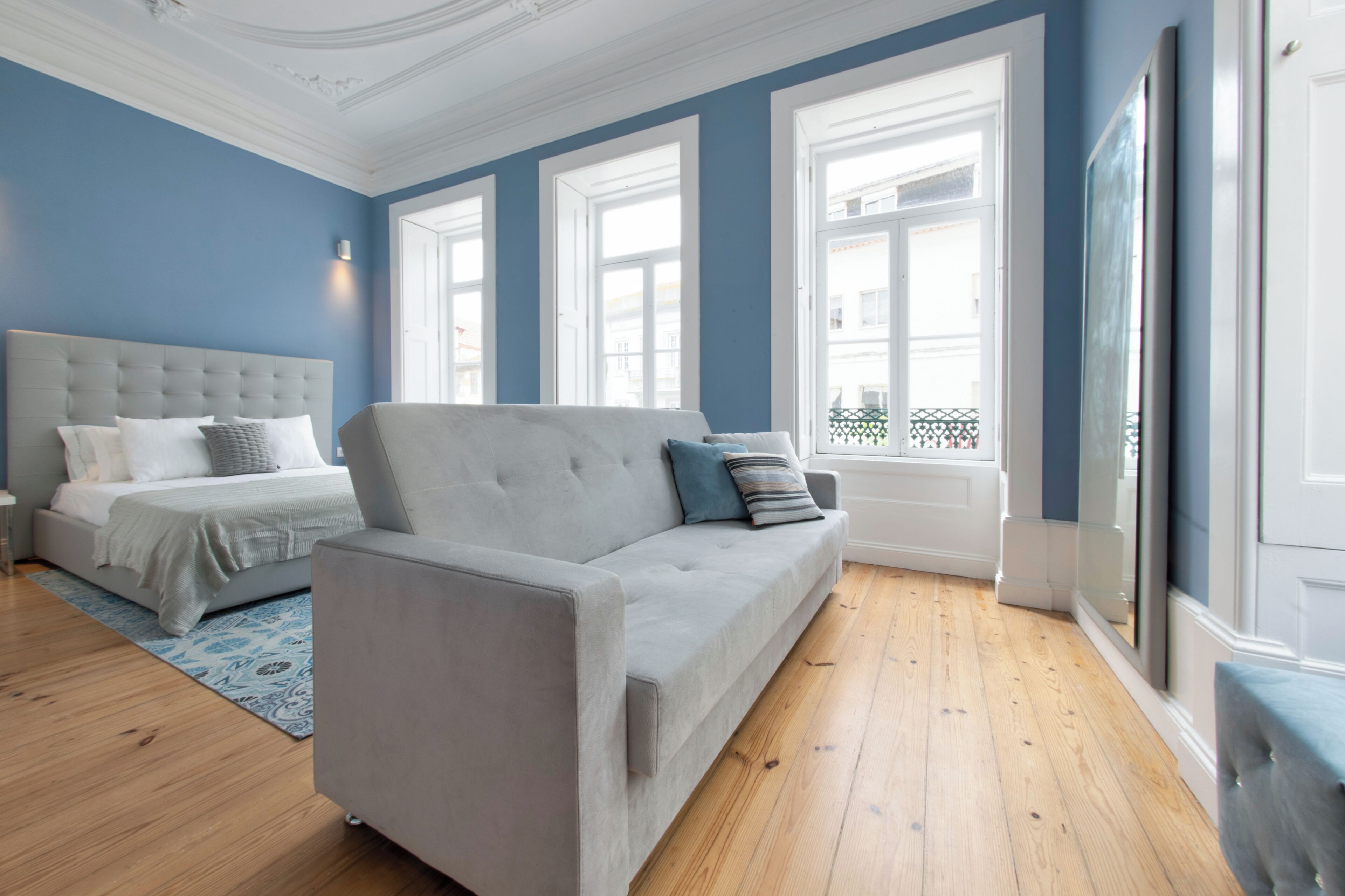 Une chambre à coucher avec un lit, un canapé gris et une fenêtre donnant sur des immeubles.