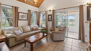 A spacious living room with a wooden ceiling, a leather sofa, a wooden coffee table, and a chair. There are large windows with curtains, a clock on the wall, and a lamp on a side table. - Private Coastal 4 BD Getaway in Ocean Park ME (Old Orchard Beach)