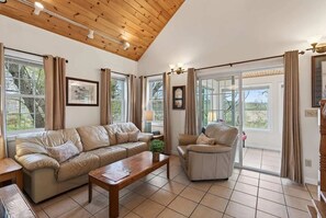 A spacious living room with a wooden ceiling, a leather sofa, a wooden coffee table, and a chair. There are large windows with curtains, a clock on the wall, and a lamp on a side table. - Private Coastal 4 BD Getaway in Ocean Park ME (Old Orchard Beach)
