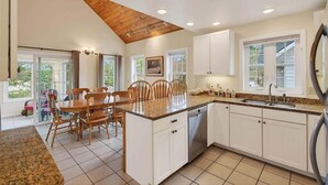 A kitchen with white cabinets, a granite countertop, a wooden dining table with chairs, and a view through the glass door. - Private Coastal 4 BD Getaway in Ocean Park ME (Old Orchard Beach)