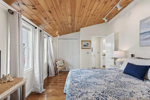 Ein Schlafzimmer mit Holzboden, einem Bett mit gemustertem Überwurf, einem Sessel, einem Nachttisch und einem Bild an der Wand.