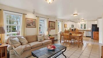 A spacious living room with a sofa, dining table, and chairs, a kitchen area with cabinets and appliances, and large windows with curtains.
