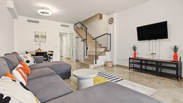 A modern living room with a grey sectional sofa, a round coffee table, a staircase, a flat-screen TV, and a dining area with a table and chairs.