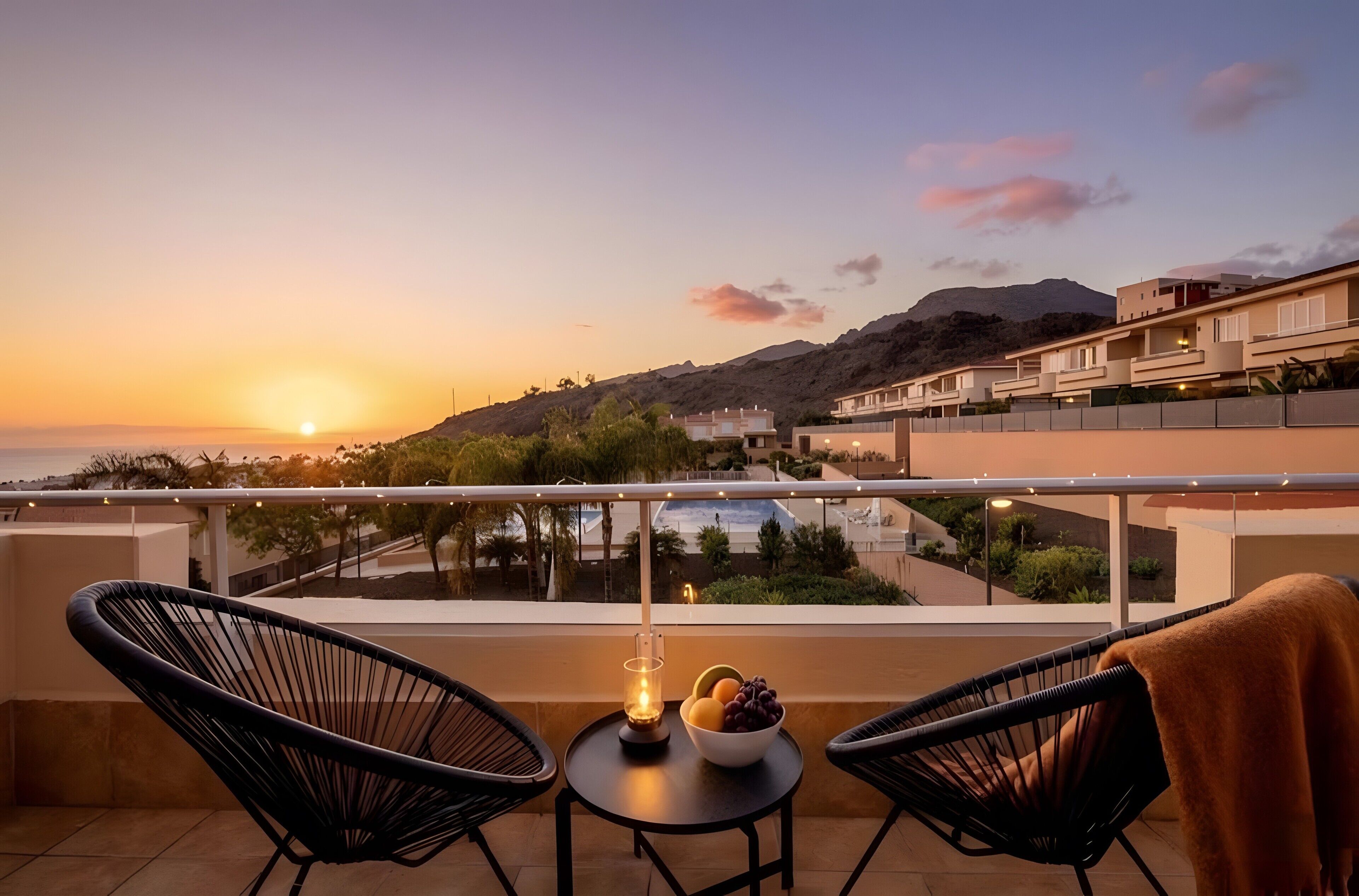 A balcony with two chairs, a table with fruit, and a sunset view.