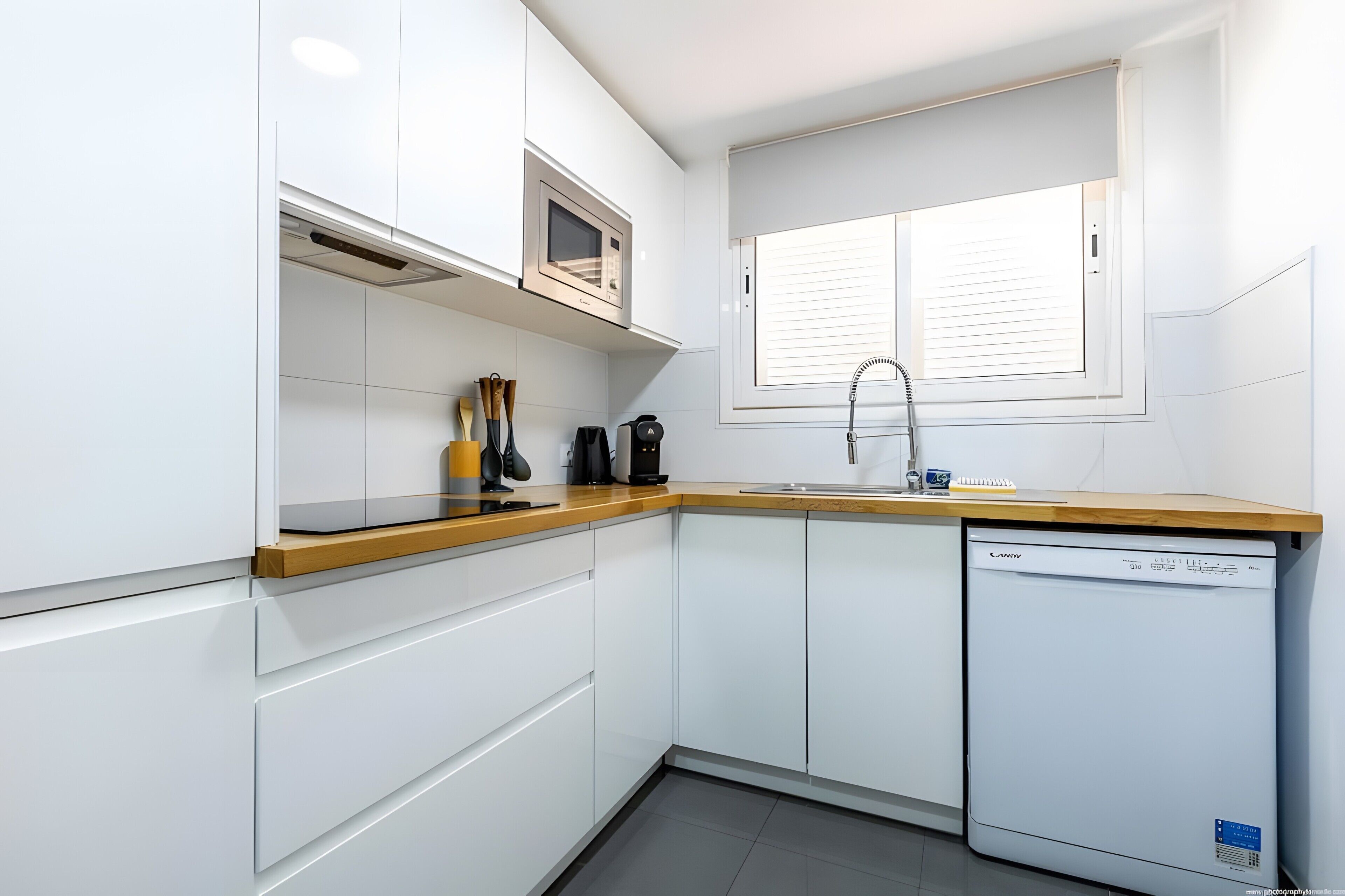 A modern kitchen with white cabinets, a built-in microwave, a dishwasher, and a window with blinds.
