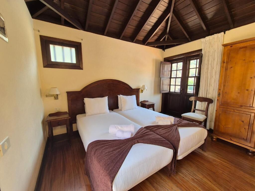 A double bed with a wooden headboard, two bedside tables, a nightstand, a window with curtains, and a wooden wardrobe.