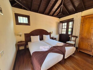 Uma cama de casal com cabeceira de madeira, duas mesas de cabeceira, um criado-mudo, uma janela com cortinas e um guarda-roupa de madeira.