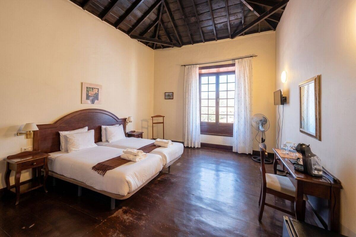 Habitación de hotel con dos camas, techo de madera, ventana con cortinas, escritorio con ventilador y una silla.