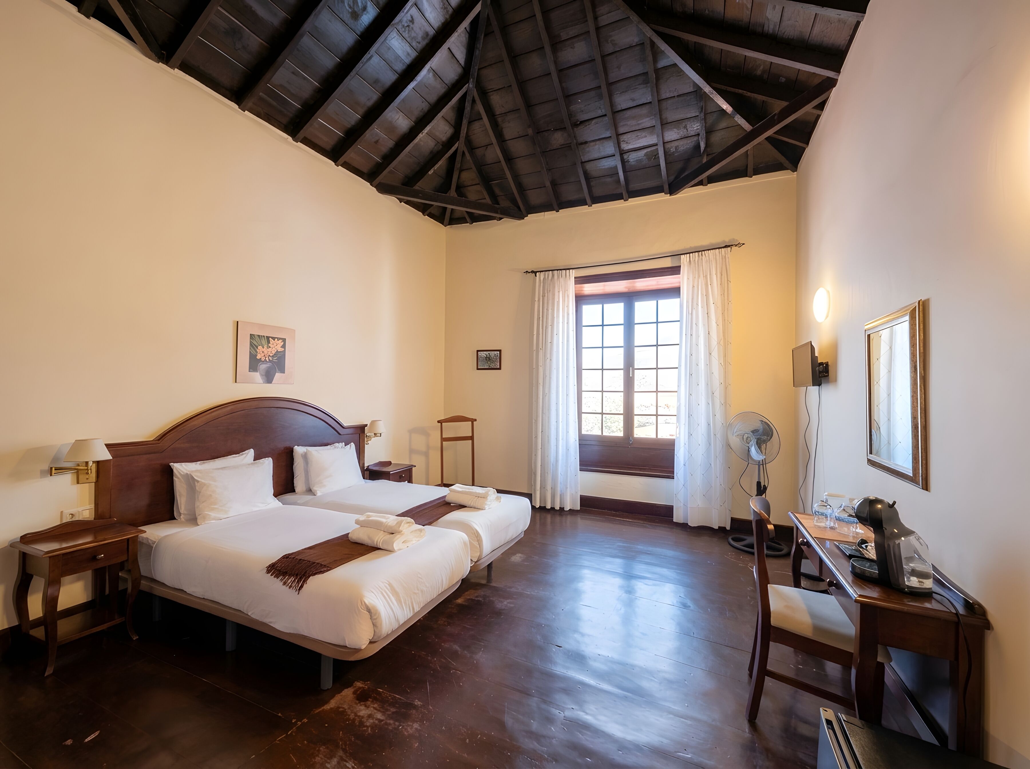 A hotel room with two beds, a wooden ceiling, a window with curtains, a desk with a fan, and a mirror.