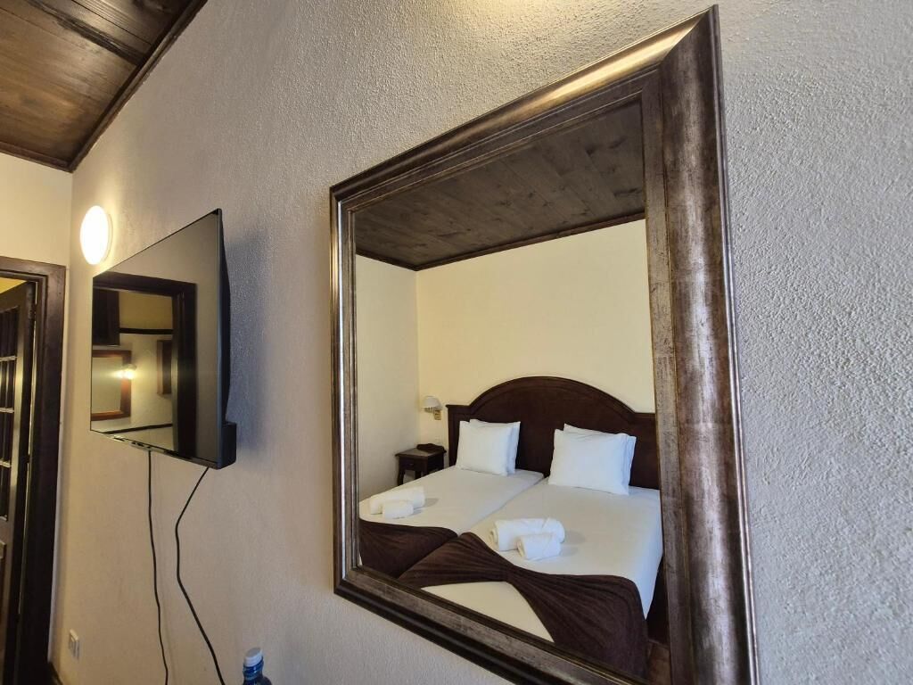 Une chambre d’hôtel avec un grand miroir, un lit avec du linge de maison blanc, une tête de lit en bois et une table de chevet avec une lampe.