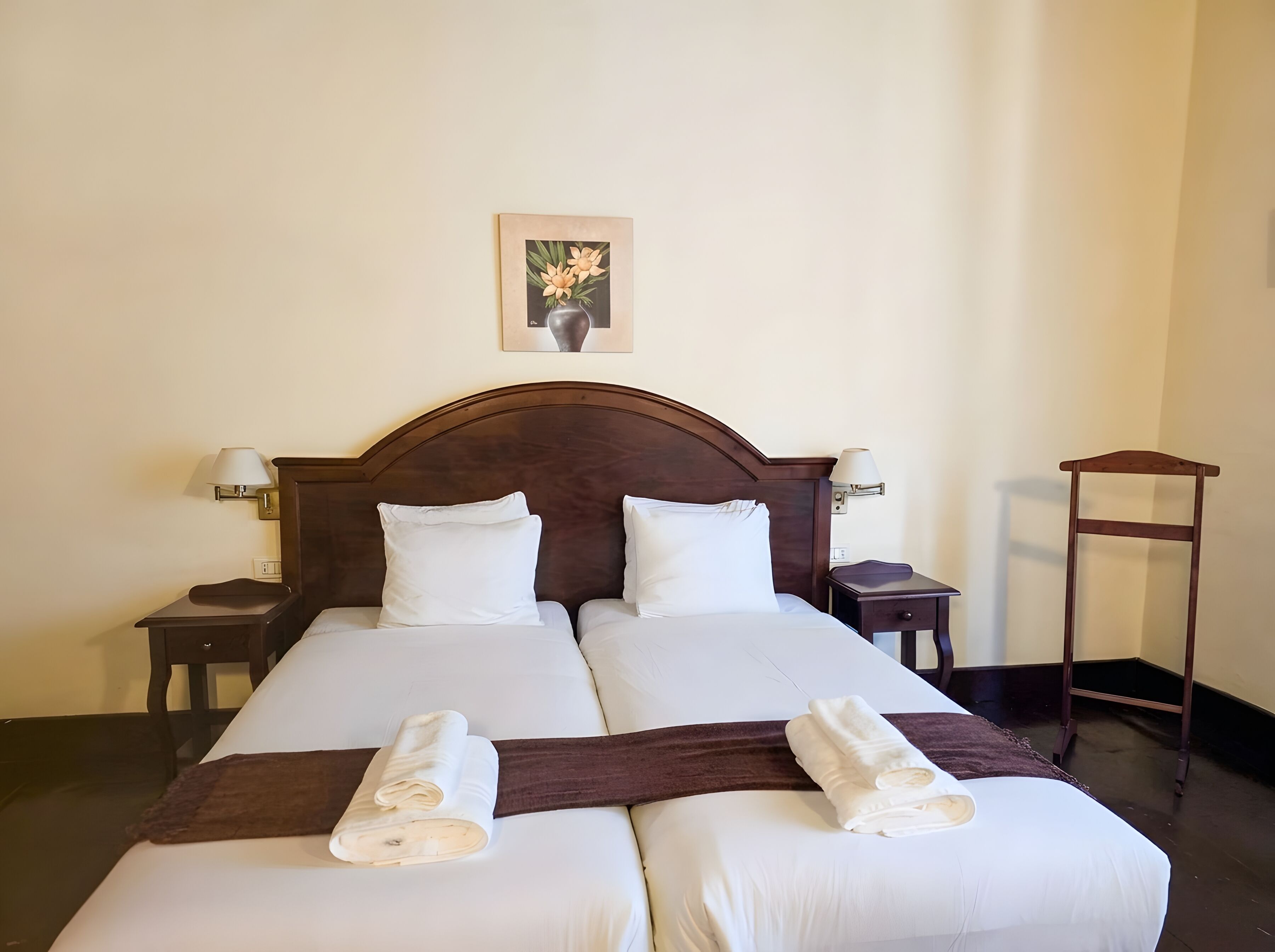 A double bed with white linens, two bedside tables, a nightstand, a wooden chair, and a framed picture on the wall.
