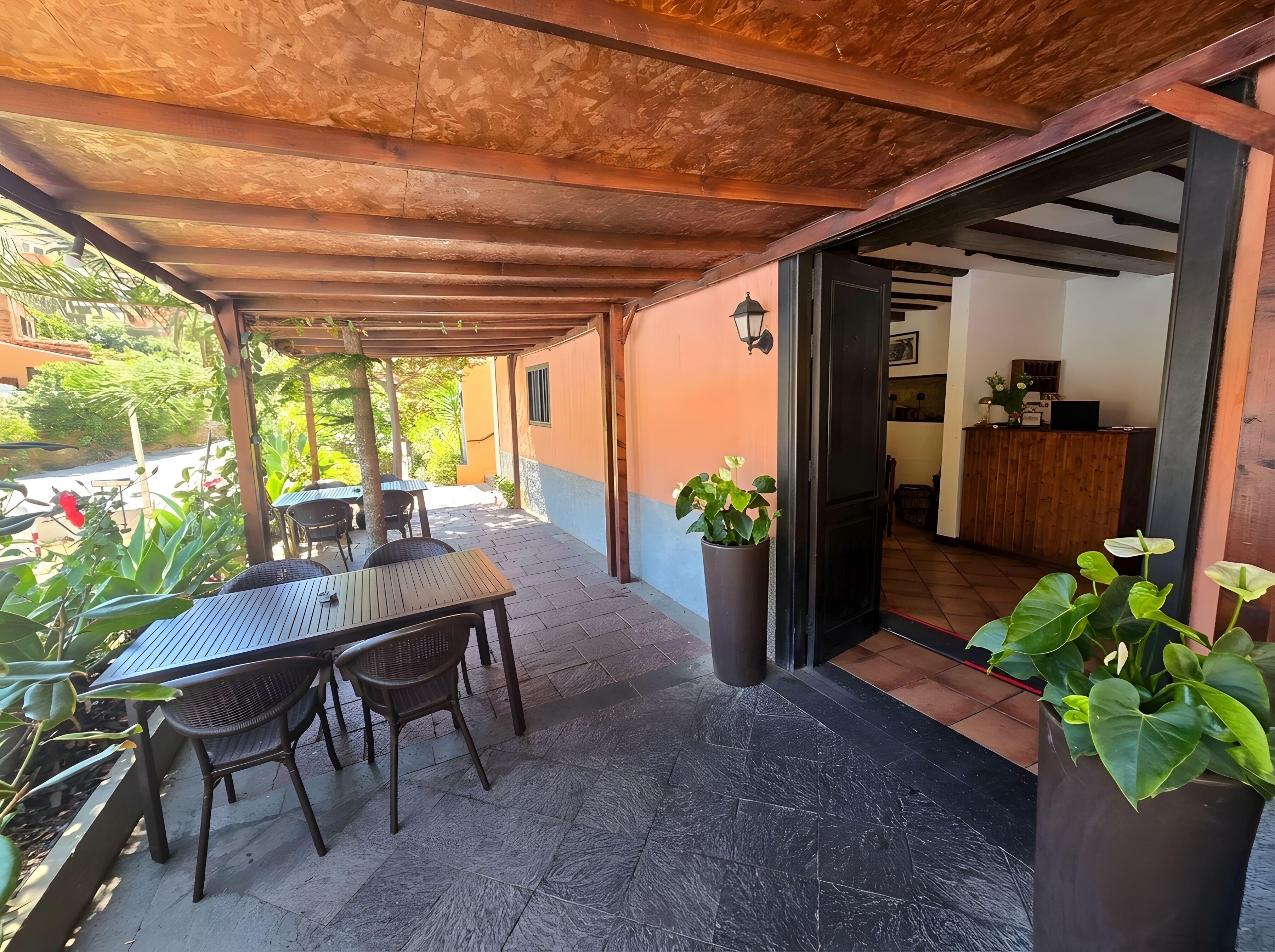 Een terras buiten met een tafel, stoelen, een houten plafond en een deur naar binnen.