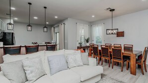 A modern living room with a sofa, dining table, chairs, pendant lights, and a kitchen area. - 5BR Private Pool Home at Veranda Palms (Kissimmee)