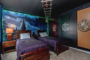 Quarto com duas camas, mesa de cabeceira, abajur, parede com um brasão e um mural do céu estrelado.