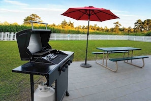 An outdoor grill with a red umbrella, a picnic table, and a white picket fence in the background.