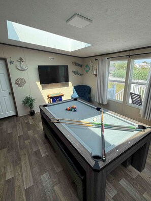 A pool table with pool cues, a television, and decorative fish wall art in a room with wooden flooring and a skylight. - Stay Under the Stars in Chincoteague (Chincoteague)