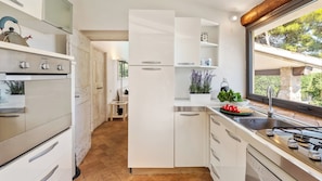 A modern kitchen with white cabinetry, a built-in oven, and a window with a view of greenery. - Villa Althaea 6, Emma Villas (Orbetello)