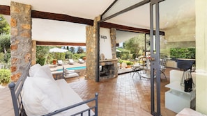 A covered outdoor seating area with stone pillars, a pool, and a barbecue grill. - Villa Althaea 6, Emma Villas (Orbetello)
