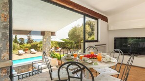 Ein Essbereich mit Blick auf einen Swimmingpool und Grünflächen, inklusive Fernseher und Tisch, gedeckt für das Frühstück.