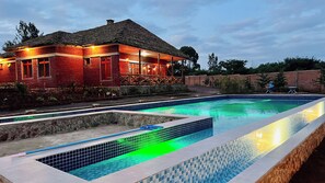 Una piscina con borde de azulejos, una casa de ladrillo con techo de paja y un muro de piedra al fondo.