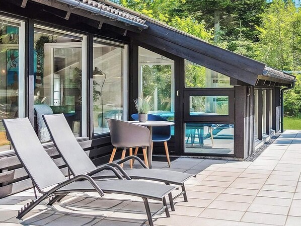 A modern outdoor seating area with lounge chairs and a table, a glass-enclosed patio, and a wooden pergola structure. - 5 Star Holiday Home in Bindslev (Bindslev)