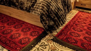 A bedroom with a stone wall, a bed with a dark blanket, a red patterned rug, and a nightstand with a lamp.