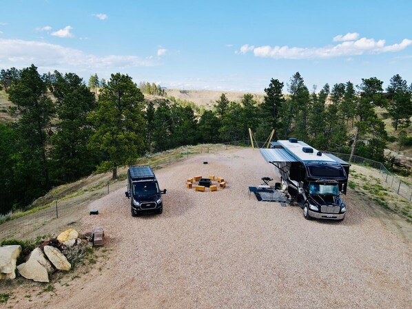 A gravel parking area with two RVs and a car, surrounded by trees and a hilly landscape.