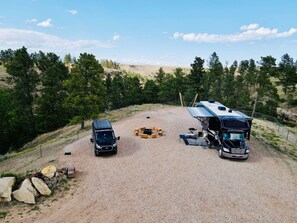 A gravel parking area with two RVs and a car, surrounded by trees and a hilly landscape.