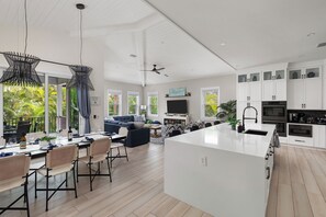 A modern kitchen with white cabinetry, a large island, and a dining area with a view of the outdoors. - Heart of Pine Ave | 6 En-Suites | Heated Pool (Anna Maria)