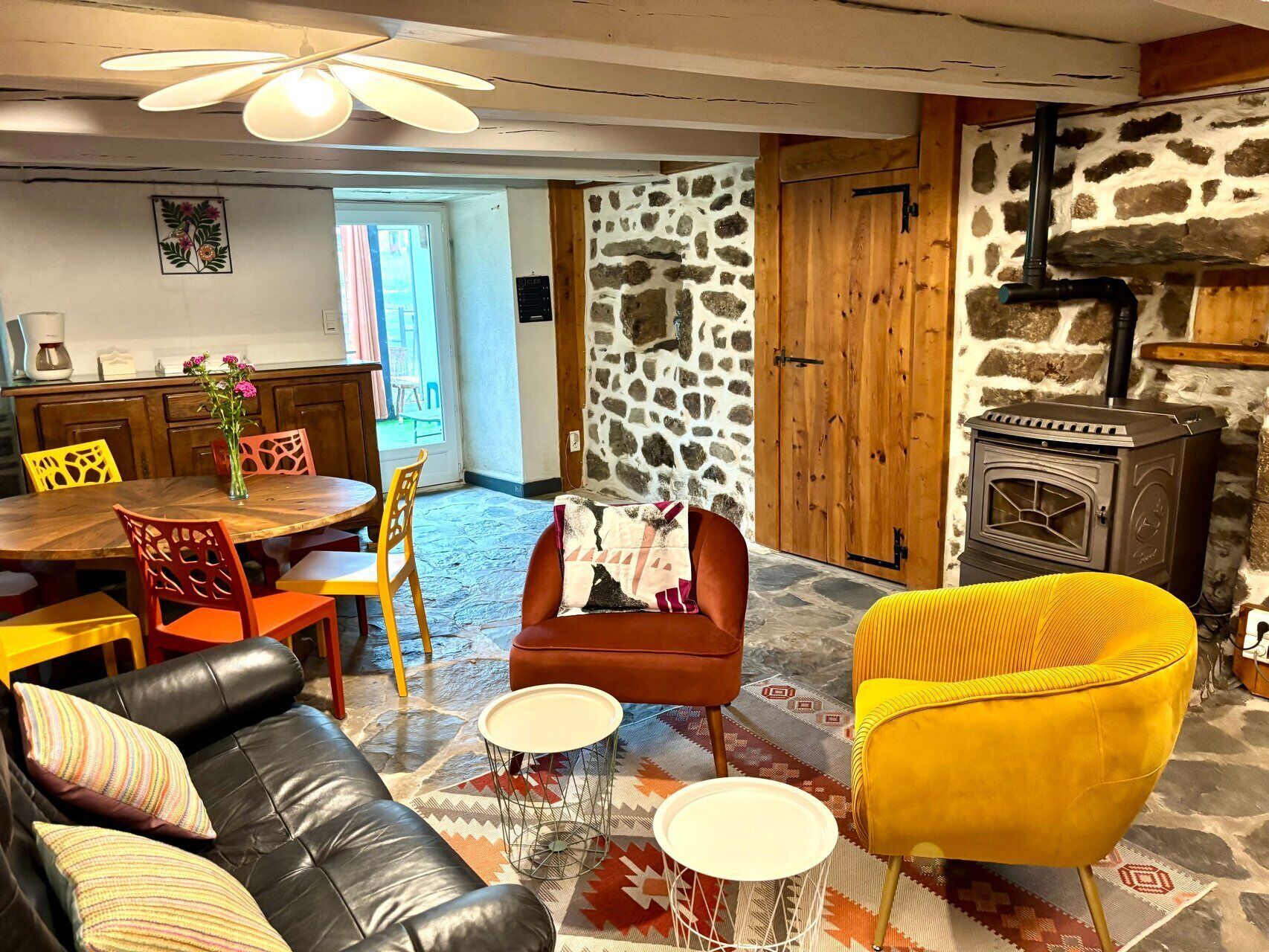 Una sala de estar acogedora con pared de piedra, chimenea, comedor con mesa y sillas, y sofá con cojines estampados.