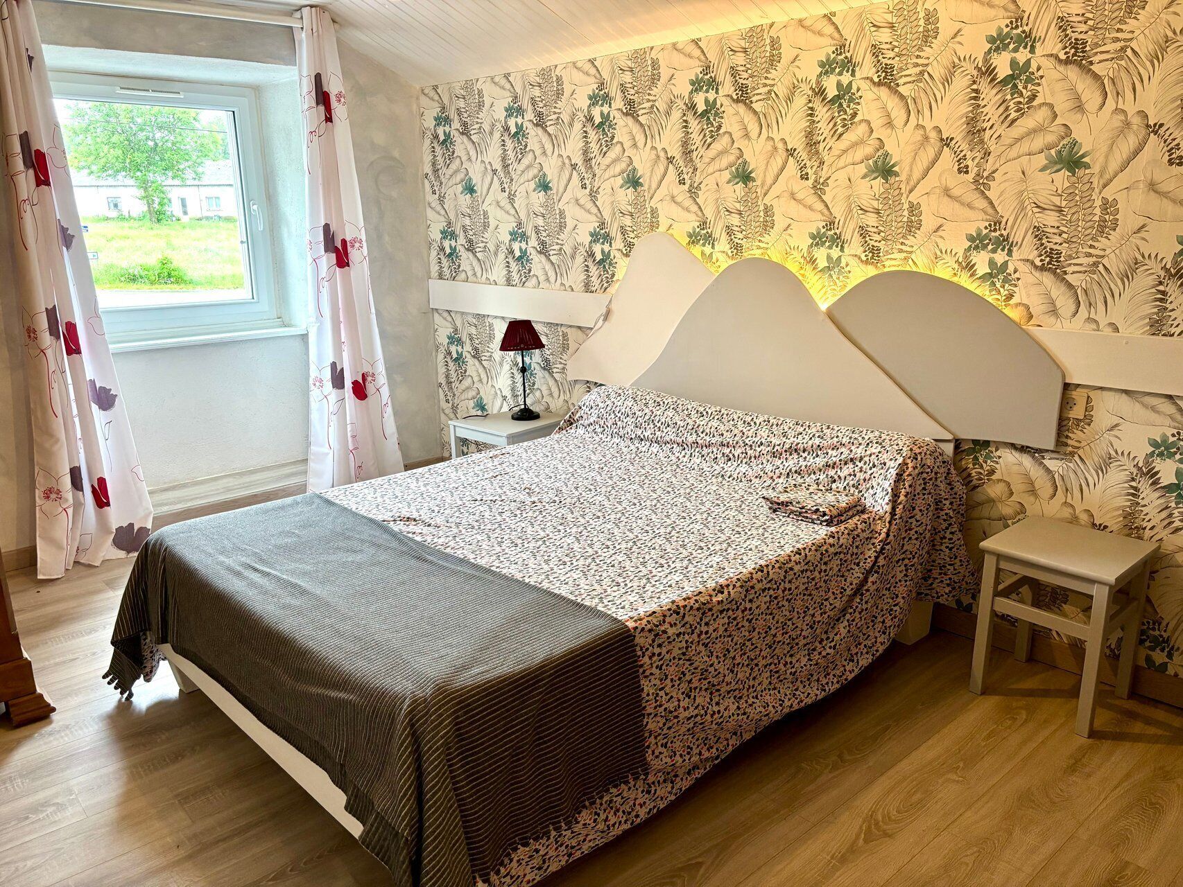 A bedroom with a bed, a nightstand, a lamp, a window with curtains, and a wall with a floral wallpaper.