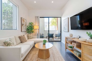 A modern living room with a sofa, coffee table, TV, and a view through a glass door to an outdoor area. - Modern Little Italy Retreat w/ Patio & Parking (San Diego)