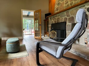 Una sala de estar con chimenea de piedra, un sillón tapizado de gris, un reposapiés redondo y suelo de madera.