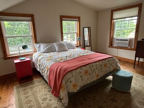 Un dormitorio con cama, dos ventanas, una mesita de noche, un espejo y un aparador.