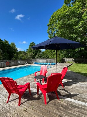 A wooden deck with a pool, red chairs, and a blue umbrella.