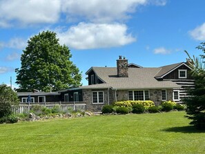 A two-story stone house with a large lawn, a wooden deck, and a prominent tree.