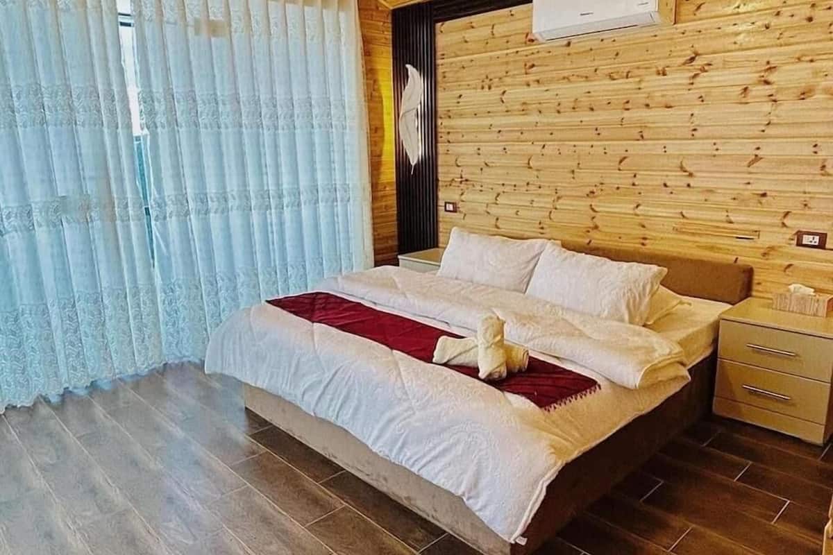 Un dormitorio con cabecera de madera, cama con ropa de cama blanca, un plaid rojo, una mesita de noche y un mueble con cajones.