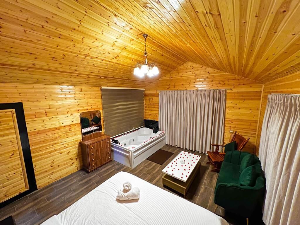 A wooden cabin interior with a bed, a sitting area, a bathtub, and a green chair.