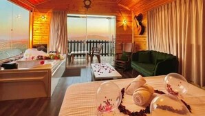Una cabaña con interior de madera, una cama con cabecera decorativa, un comedor y vistas a través de un ventanal amplio.