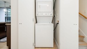 Uma lavanderia compacta com máquina de lavar e secadora, uma escada de madeira e vista para a cozinha.