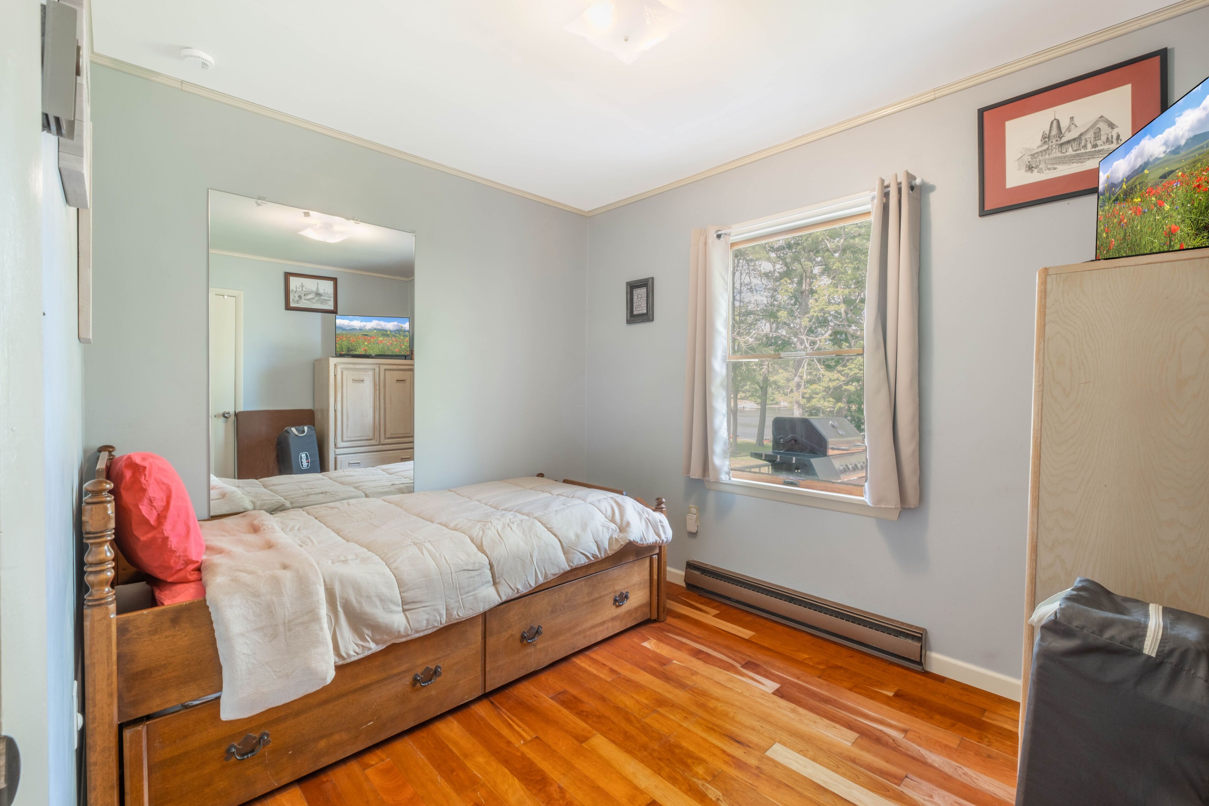 A bedroom with a wooden bed, a window with curtains, a radiator, and a mirror reflecting a door and a picture frame.