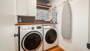 A laundry room with two washing machines, a wooden countertop, a shelf with decorative items, and a towel rack with towels.