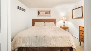 Une chambre à coucher avec un lit, une table de chevet, une lampe, une commode, un tableau encadré et un panneau accroché au mur.