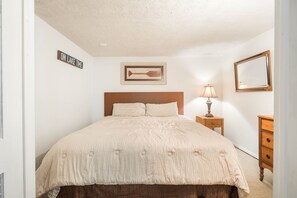 A bedroom with a bed, a nightstand, a lamp, a dresser, a framed picture, and a sign on the wall.